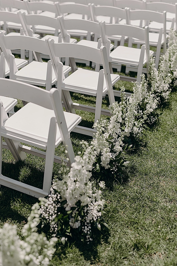 Ceremony aisle decor with outdoor ceremony aisle flowers lining a grass lawn aisle between white folding chairs with greenery accents