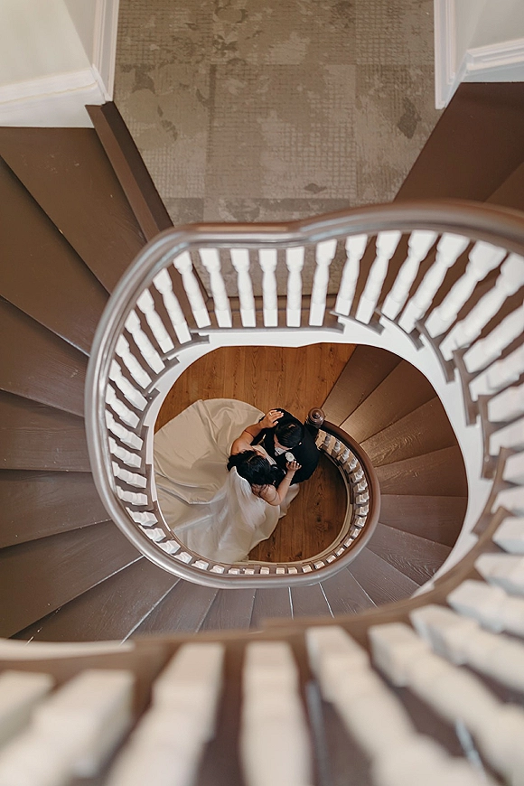 Couple portrait in an overhead wedding photo, bride in veil and gown hugging groom in black tux on a wooden spiral staircase indoors