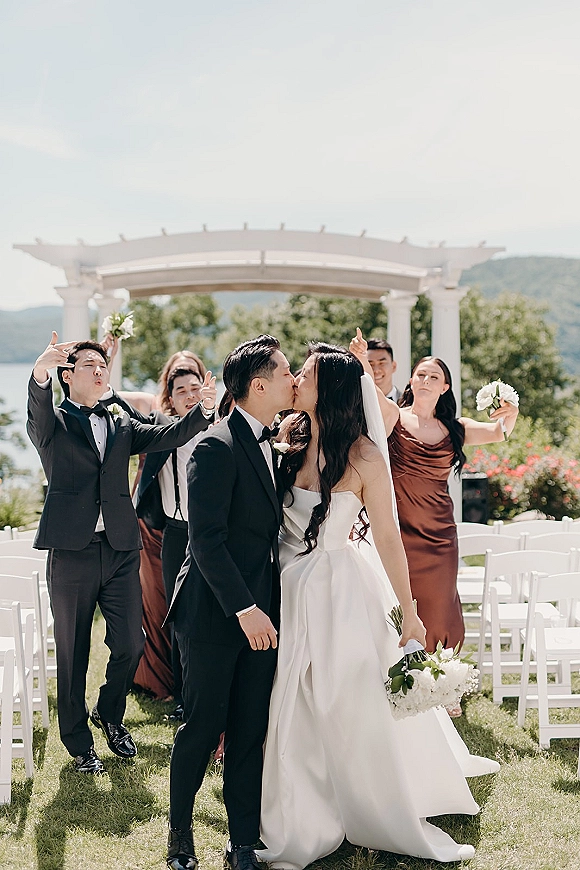 Wedding kiss portrait of bride and groom kissing as wedding party cheers, bouquet and veil visible on a lawn by lake and mountains