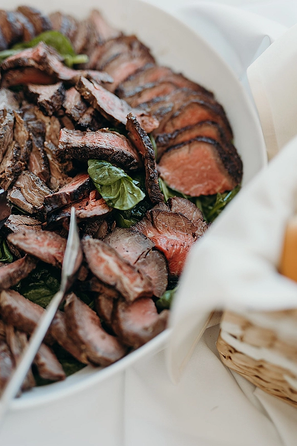 Wedding catering food platter with sliced beef, leafy greens, and serving tongs on a white tablecloth buffet table setting