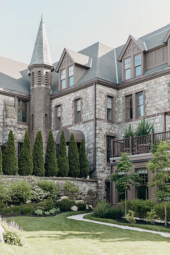Wedding venue exterior of a stone manor wedding venue with turret tower, slate roof and balcony above manicured lawn and shrubs