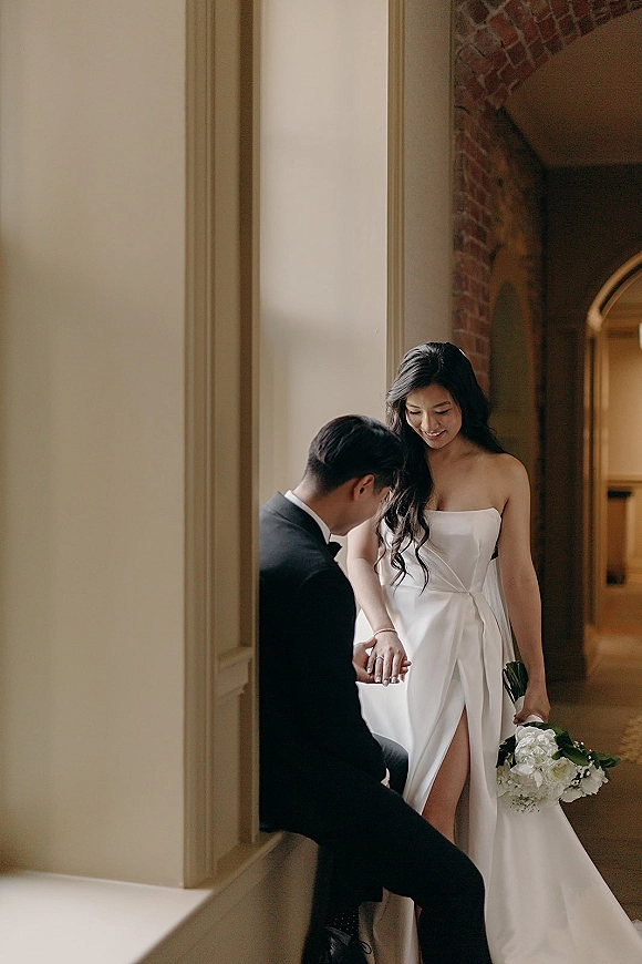Couple portrait of groom kissing the bride’s hand as they hold hands, her bouquet and strapless dress glowing in a warm brick hallway doorway