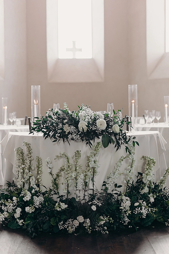 Wedding sweetheart table with white floral centerpiece and greenery garland, candlelit with tall glass cylinders, set before chapel windows and cross
