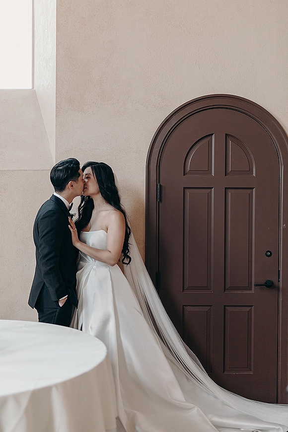 Wedding kiss portrait of bride and groom kissing in doorway, her strapless gown and long veil lit by window light against stucco wall
