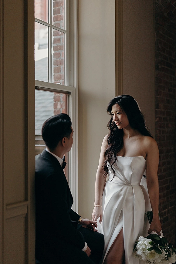 Couple portrait of bride and groom by window, bride in strapless satin dress holding bouquet beside brick wall in soft natural light