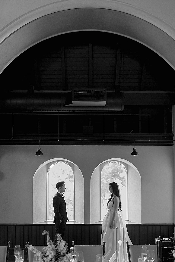 Couple portrait of bride and groom facing each other beside arched windows, with reception table candles and floral centerpiece in foreground