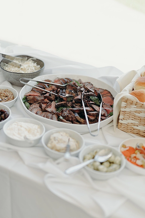 Wedding buffet food display with sliced roast beef platter, serving tongs, white bowls of sauces, and bread rolls on a white tablecloth buffet table