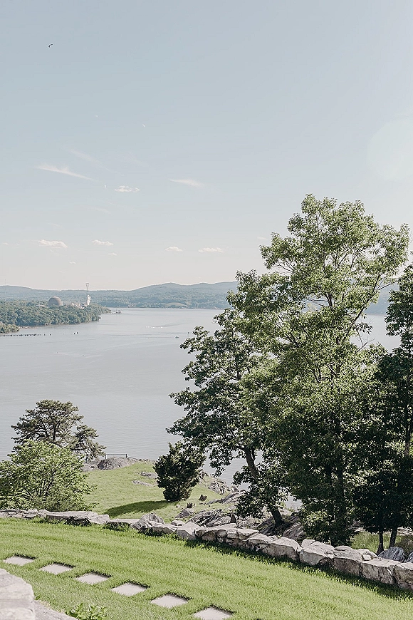 Lakeside venue view with a stone wall accent, stepping stones across a grassy lawn leading to a lake framed by trees and hills