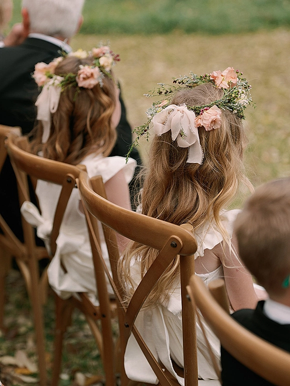 Flower girls in white dresses and flower girl floral crowns with ribbon bows sit on wooden chairs during an outdoor grass field ceremony