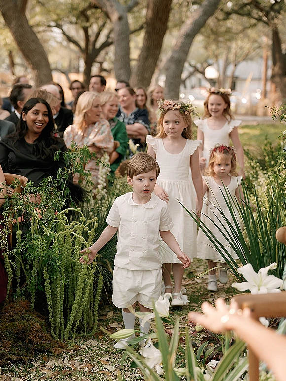 Flower girl and ring bearer walk the aisle with a flower crown and white outfits, passing greenery and lilies at an outdoor garden ceremony