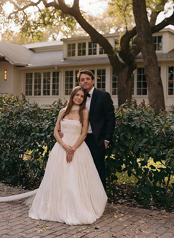 Couple portrait of bride in strapless gown and veil with groom in black tuxedo and bow tie on a brick garden walkway by a house