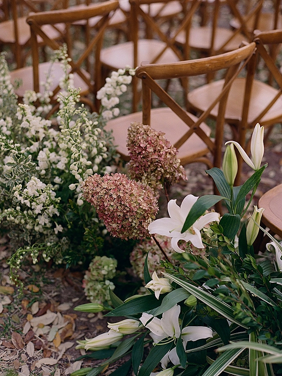 Ceremony aisle florals with wedding aisle flowers in white lilies and hydrangea clusters along a gravel path beside wooden cross-back chairs