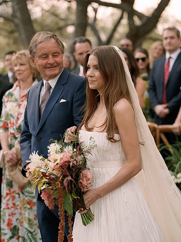 Wedding processional as bride walking down aisle with her father, holding a blush bouquet and veil flowing past wooden chairs in a garden setting