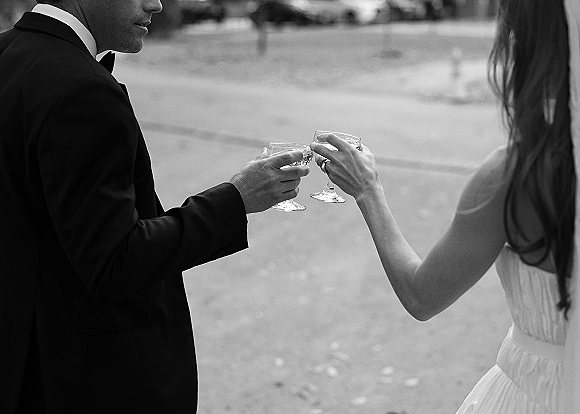 Wedding toast as bride and groom clink champagne coupes, showing wedding rings and tuxedo bow tie on an outdoor lawn with trees