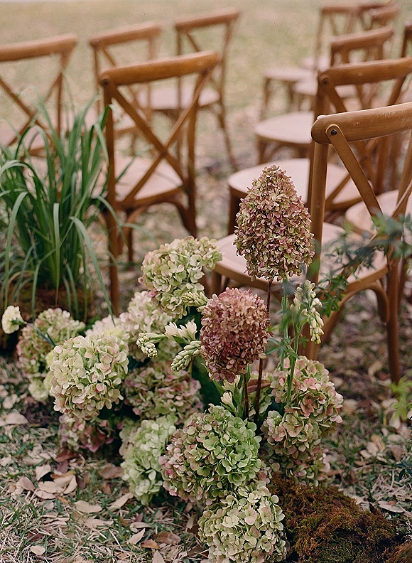 Ceremony aisle decor with outdoor ceremony chairs, featuring low hydrangea and greenery clusters on moss beside wooden crossback rows on grass lawn