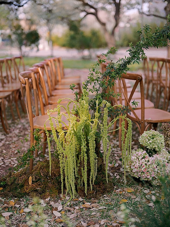 Ceremony aisle decor with outdoor ceremony chairs lined beside moss, hydrangea and hanging greenery on a garden lawn under trees