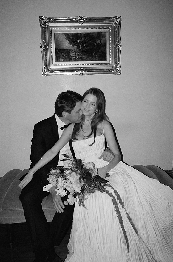 Wedding couple portrait of groom kissing bride’s cheek as they sit on a sofa, bride holding bouquet with greenery by ornate framed artwork