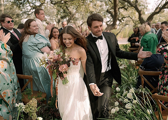 Wedding recessional as newlyweds walking down aisle, bride in strapless dress holding bouquet, guests cheering in garden setting