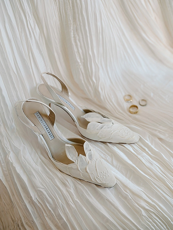 Wedding shoes in a bridal slingback heels flat lay with floral applique and gold wedding bands on pleated white fabric over wood floor