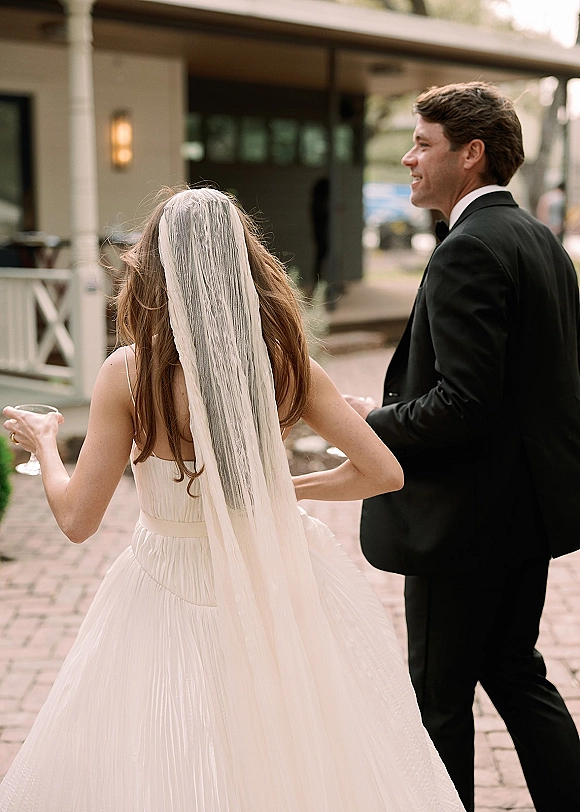 Wedding couple portrait of bride and groom walking with champagne coupes, her long veil over a strapless dress on a brick patio walkway