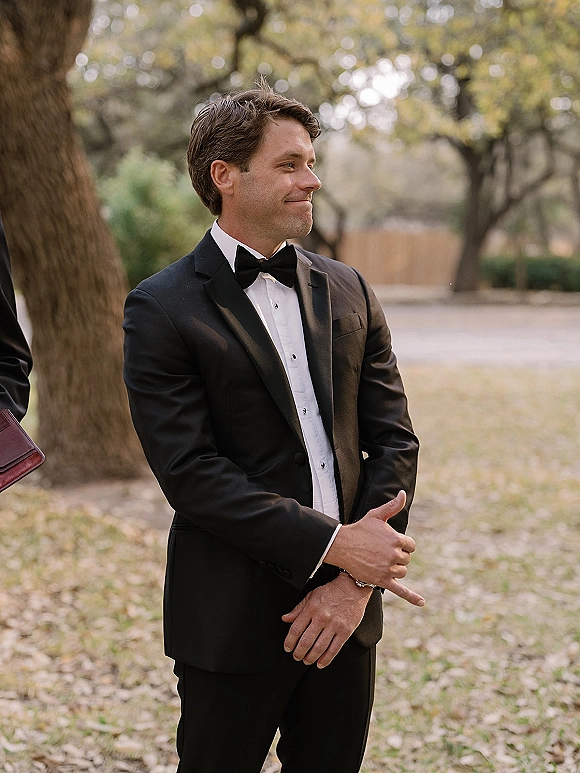 Groom portrait in a black tuxedo with bow tie, cufflinks, and wristwatch, standing on an outdoor path with trees and fallen leaves