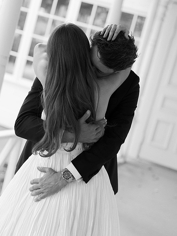 Wedding hug as bride in strapless gown embraces groom in black suit, his wristwatch visible, on a porch by window panes