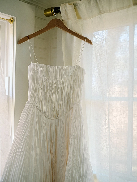 Wedding dress hanging on a wooden hanger by a sunlit window, white strapless ruched fabric with spaghetti straps and sheer curtains behind
