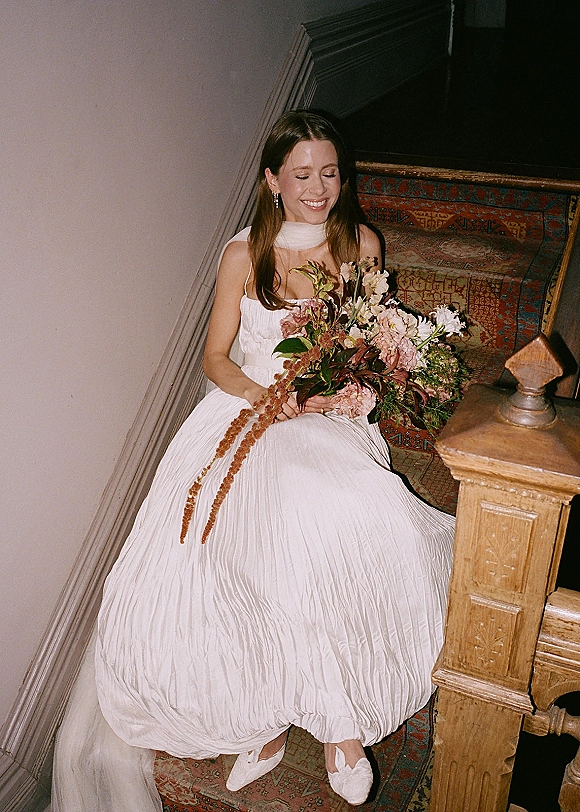 Bridal portrait of a bride sitting on stairs, smiling in a white wedding dress and holding a greenery bouquet by a patterned runner
