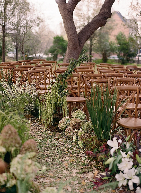 Ceremony aisle decor with wood crossback chairs and hydrangea-lined greenery garland bordering a grass runner in an outdoor garden setting