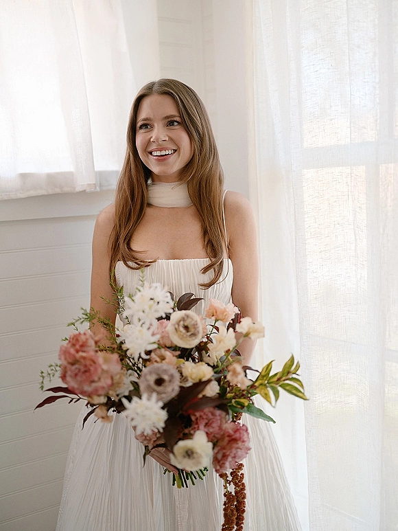 Bridal portrait of a smiling bride holding bouquet in a strapless dress and neck scarf, long hair by sheer curtains and window light