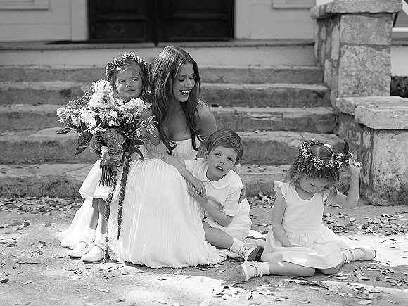 Bride with flower girls sits on stone steps holding a bridal bouquet, children in flower crowns and dresses, fallen leaves nearby