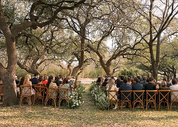 Outdoor wedding ceremony with garden wedding ceremony seating, wood cross-back chairs, and white aisle florals under oak trees on a lawn