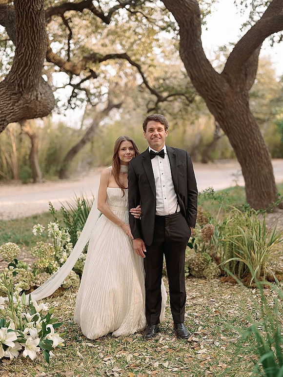 Couple portrait of bride in strapless gown and veil holding hands with groom in black tuxedo on a wooded path with oak trees and wildflowers