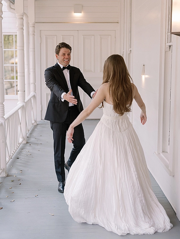 First look moment as bride in a strapless wedding dress walks up behind groom in tuxedo with arms open on a white porch with columns