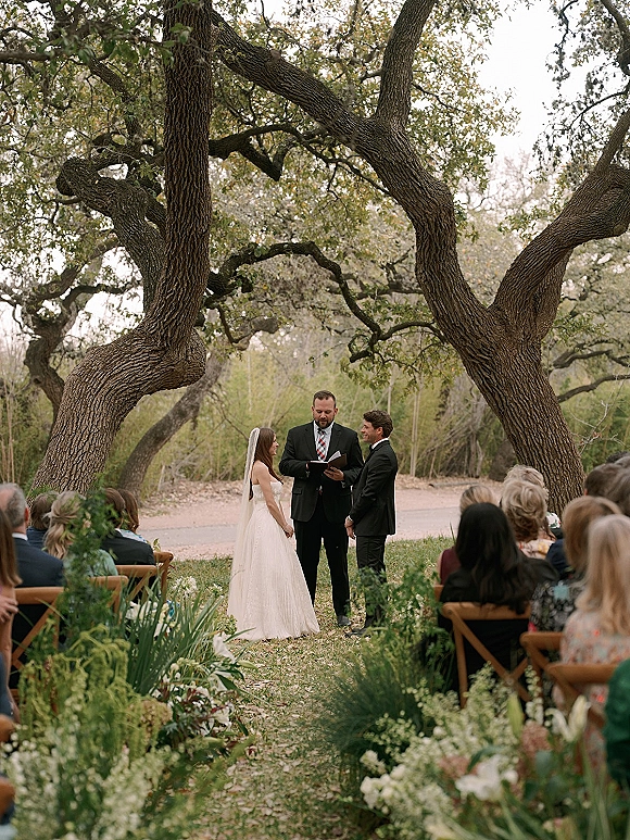 Wedding ceremony with bride and groom under oak trees, officiant reading from a book, veil trailing beside aisle florals on a lawn