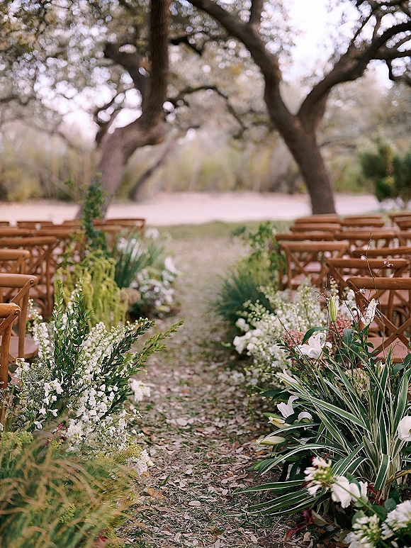Ceremony aisle decor with white lilies and greenery forming a ground floral meadow beside wooden crossback chairs under oak trees