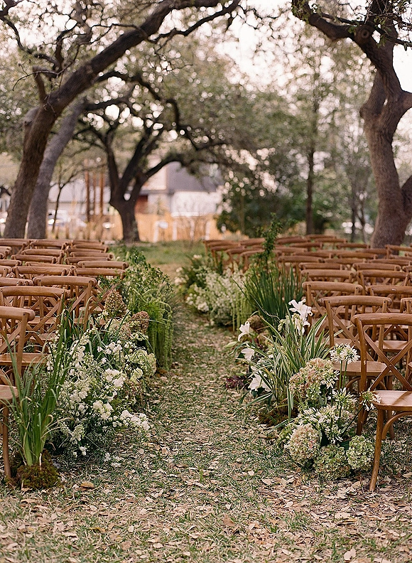 Ceremony aisle decor with outdoor wedding ceremony setup featuring low white flowers and greenery on leaf-covered grass beneath oak trees