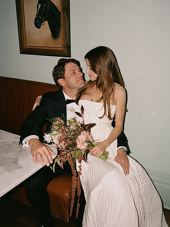 Couple portrait of bride sitting on groom’s lap in a restaurant booth, holding a bouquet in strapless dress and black tuxedo by marble table