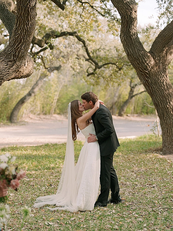 Wedding kiss portrait of bride and groom kissing, her long veil blowing as they embrace on a leaf-strewn lawn under oak trees