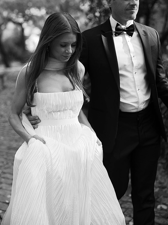 Couple portrait in a black and white wedding portrait, bride in strapless dress and groom in tuxedo walking on an outdoor path by trees
