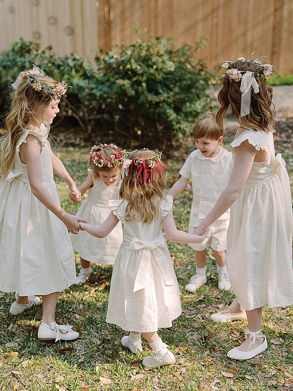 Flower girl moment as children in white dresses hold hands in a circle, wearing flower crowns on a grassy lawn by a wooden fence