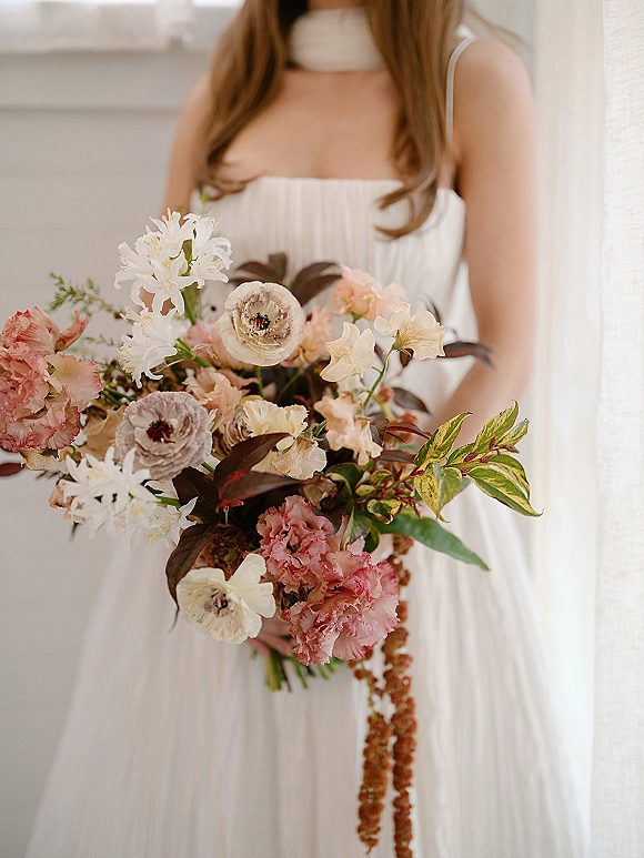 Bridal bouquet of pastel wedding bouquet blooms with greenery and a trailing floral tassel held by a bride in a strapless dress, bright window-lit room