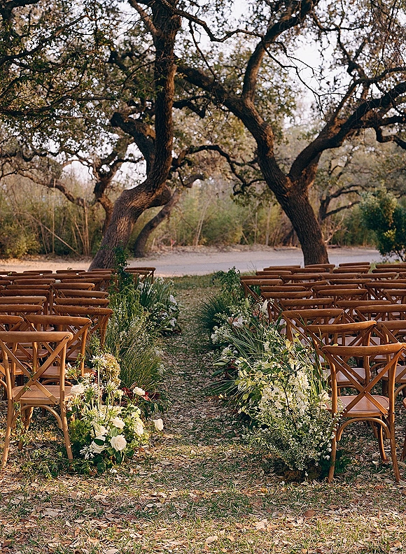 Ceremony aisle design with outdoor ceremony aisle lined in low white rose and greenery florals, flanked by wood crossback chairs under oak trees