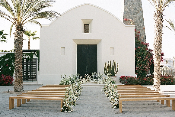 Ceremony setup with outdoor ceremony seating, wood benches lining a white-flower and greenery aisle toward a white chapel facade with black doors