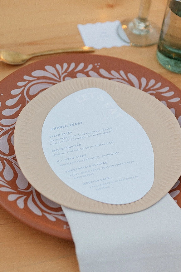 Wedding place setting with wedding menu card on a dinner plate atop a patterned charger, neatly folded napkin, flatware, and glasses on a wood table