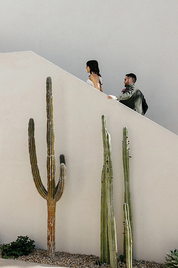 Wedding couple portrait of bride in a strapless wedding dress leading groom in sunglasses up an outdoor staircase beside tall cacti and white stucco wall