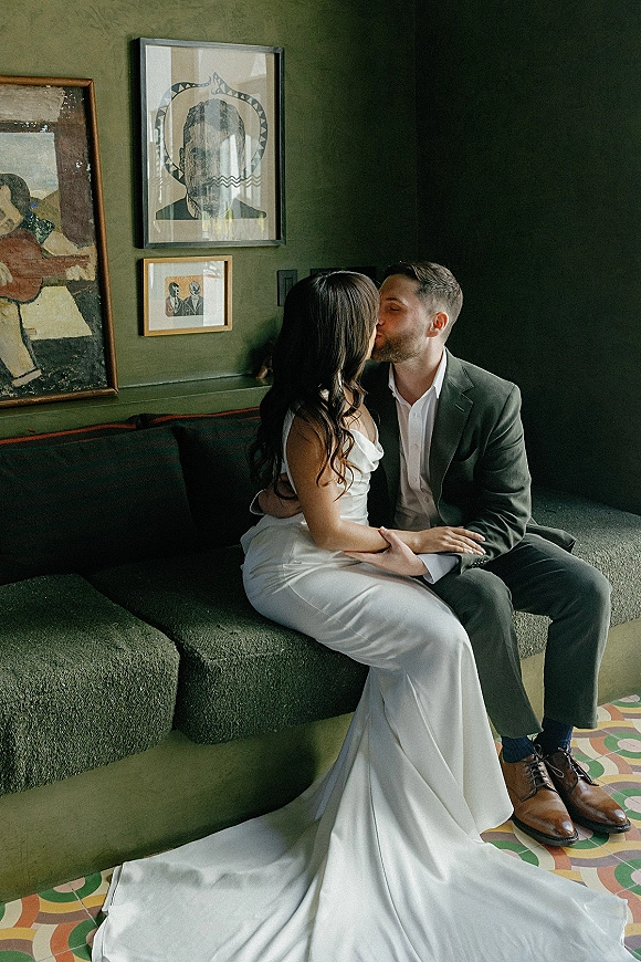 Wedding kiss portrait of bride and groom kissing on banquette, bride in strapless satin dress with train against a green wall with artwork