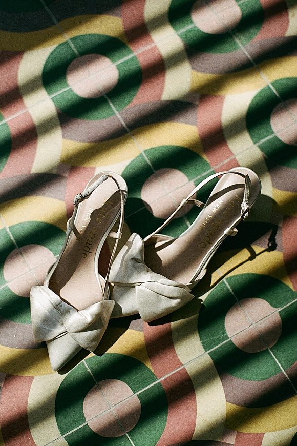 Bridal shoes in a wedding shoe flatlay with satin slingback straps and bow heels on patterned tile floor with sunlit shadows