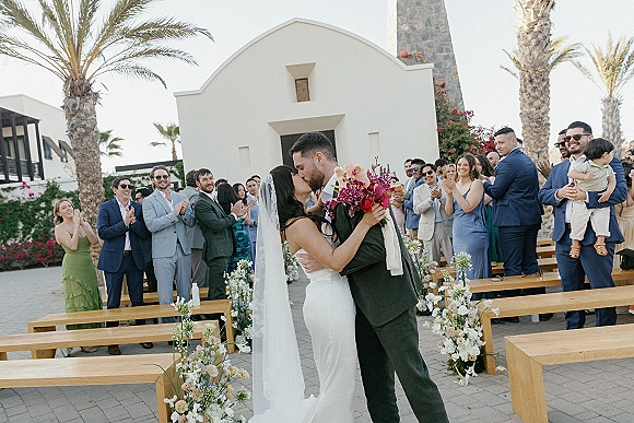 Wedding kiss at outdoor courtyard ceremony, bride in strapless dress with veil and bouquet as guests cheer by white chapel facade