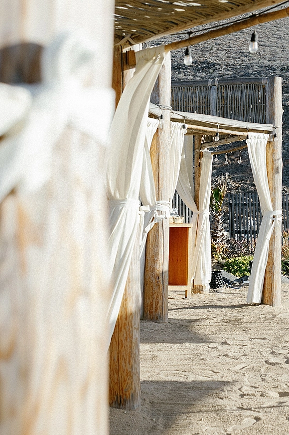 Outdoor lounge cabana wedding lounge area with white drapery on wooden posts and string lights, set on sand with hillside fence behind